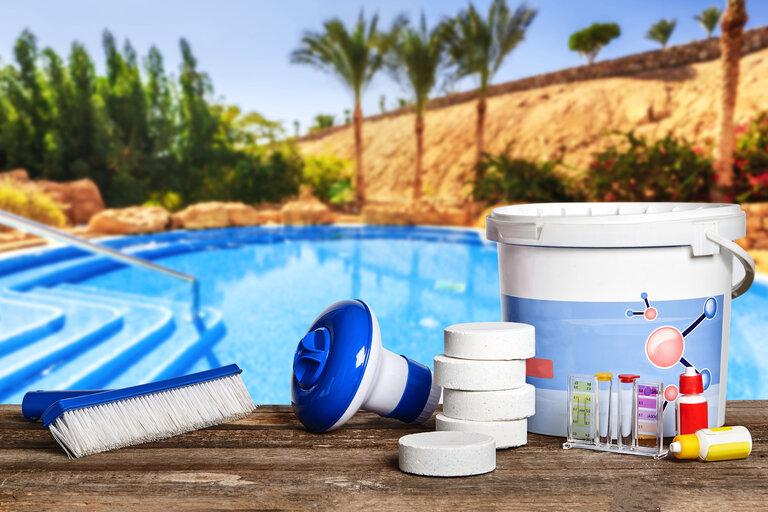Ausrüstung mit chemischen Reinigungsmitteln und Werkzeugen für die Wartung des Swimmingpools auf hölzerner Oberfläche vor einem transparenten Wasserhintergrund.